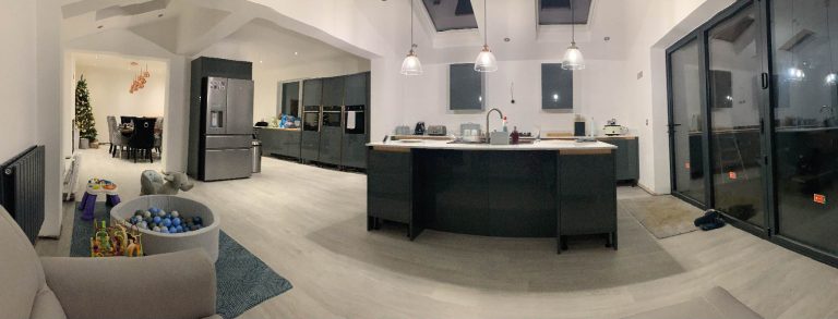 A wide angle photograph of a completed extension. On the right is a large modern kitchen with floor to ceiling windows and a glass door leading to the garden. Skylights and modern hanging light fittings provide even more light into the space. On the left is a large dining area with table and chairs.