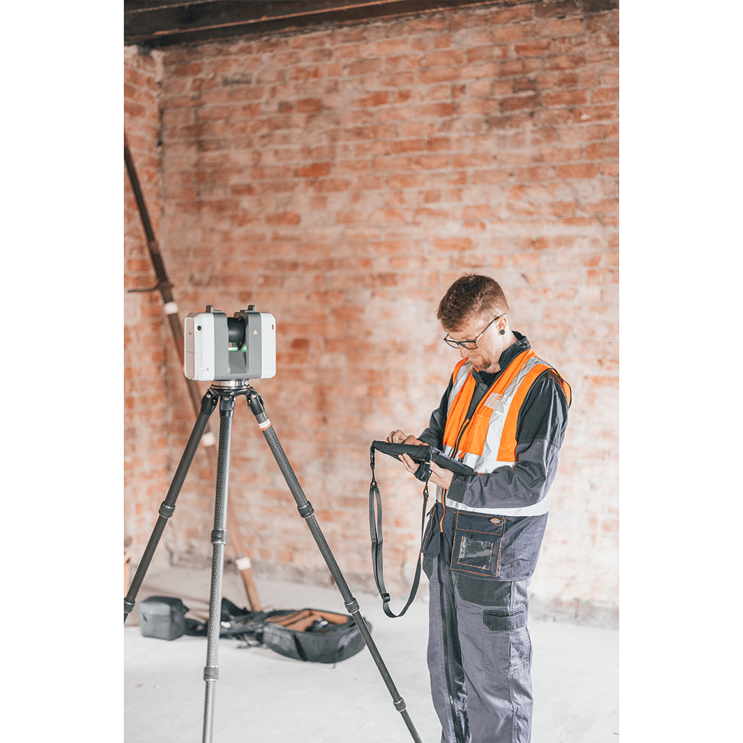 A man dressed in workwear and a high vis jacket stands looking at a tablet. To his left is a LiDAR scanner, exposed brick in the background.
